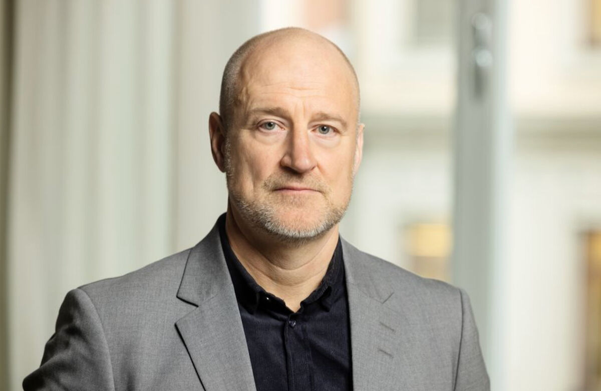 Göran Olsson er avbildet i et portrett innendørs, iført lys grå blazer og mørk skjorte. Han ser rett i kamera med et rolig, alvorlig uttrykk mot en lys og uskarp bakgrunn.