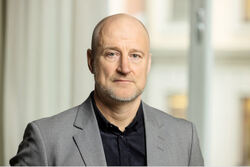 Göran Olsson er avbildet i et portrett innendørs, iført lys grå blazer og mørk skjorte. Han ser rett i kamera med et rolig, alvorlig uttrykk mot en lys og uskarp bakgrunn.