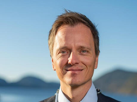 Portrett av Mads Drange i dress, fotografert utendørs i sollys. I bakgrunnen skimtes fjord og fjell under blå himmel.