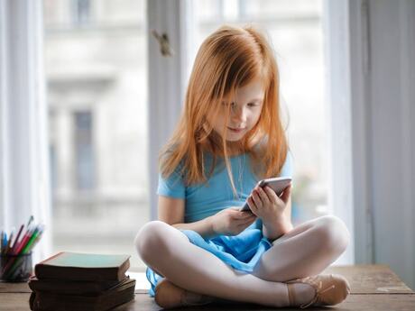 Liten jente med rødt langt hår sitter i vinduskarmen med en mobil.