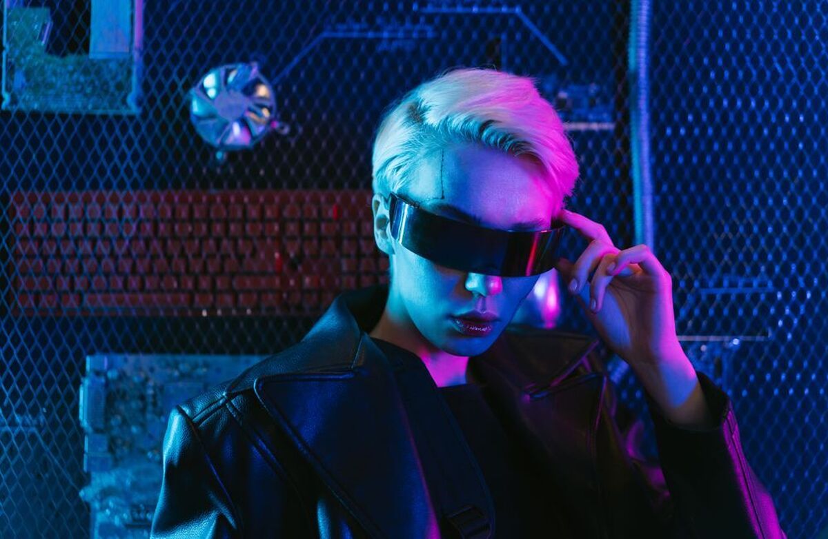 Person med lyst hår og futuristiske, mørke briller i et miljø opplyst av blått og rosa neonlys. Bildet har et tydelig cyberpunk-preg.