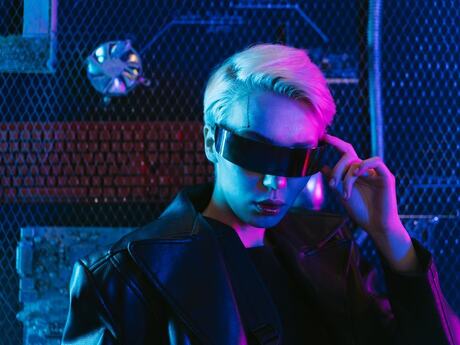 Person med lyst hår og futuristiske, mørke briller i et miljø opplyst av blått og rosa neonlys. Bildet har et tydelig cyberpunk-preg.
