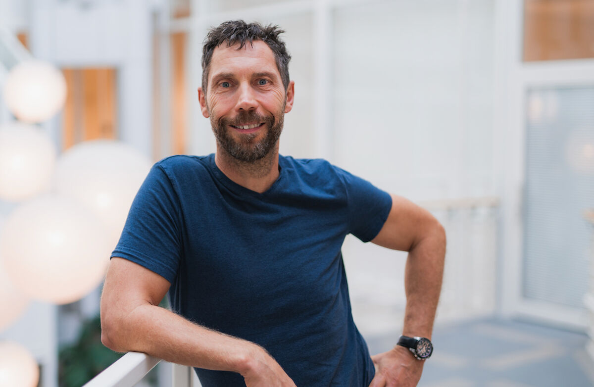 Professor Tom Karp mener norske ledere er for naive når det gjelder makt – og at tiden vi lever i krever sterkere og mer bevisste ledere. Foto: Kristiania høyskole Tom Karp smiler til kamera i lyse omgivelser, iført mørk blå T-skjorte. Han lener seg avslappet mot et rekkverk med armen hvilende på siden.