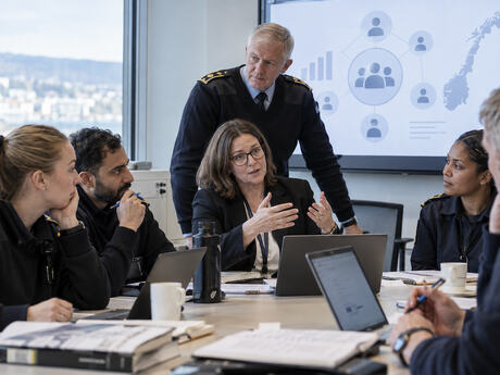 Et team av politifolk og en sivil leder sitter i møte rundt et bord med PC-er og papirer, mens en skjerm i bakgrunnen viser organisasjonsgrafikk og et kart over Norge.