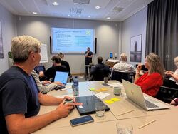 Bildet viser deltakere på en arbeidsøkt i SAMT-BU-prosjektet på Gardermoen i august 2026. Representanter fra ulike offentlige virksomheter er samlet for å dele erfaringer og utvikle mer sammenhengende tjenester for barn og unge. På skjermen presenter