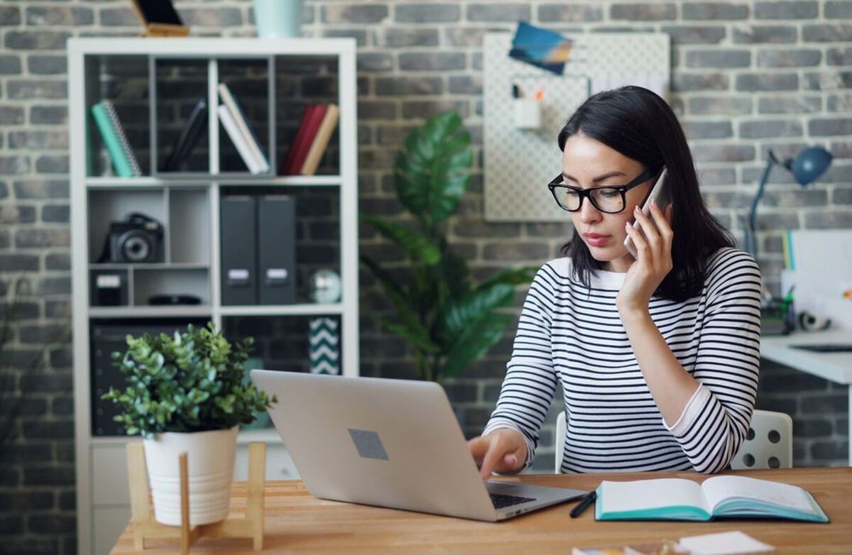 Bildet viser en kvinne som arbeider ved en PC og snakker i telefonen. Det passer godt til artikkelens tema om digital sikkerhet i en travel arbeidshverdag.