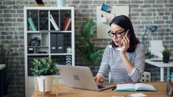 Bildet viser en kvinne som arbeider ved en PC og snakker i telefonen. Det passer godt til artikkelens tema om digital sikkerhet i en travel arbeidshverdag.