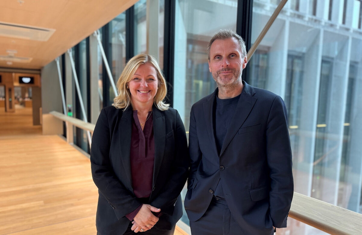 Arbeids- og inkluderingsminister Kjersti Stenseng og digitaliserings- og forvaltningsminister Torgeir Micaelsen vil ha mer inkludering og økt bruk av kunstig intelligens i staten fram mot 2029. Foto: DFD. Kjersti Stenseng og Torgeir Micaelsen står side om side i en lys, moderne korridor med store glassvinduer. Begge smiler mot kameraet og er kledd i mørke jakker.